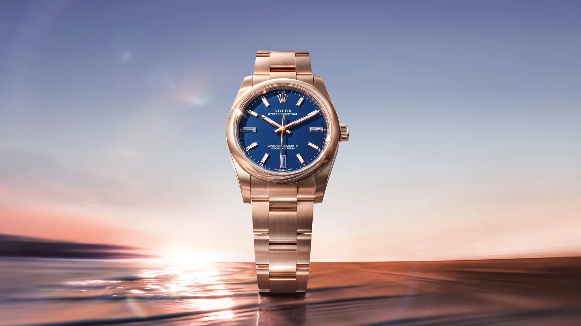 Ortada, deniz kenarında gün batımı manzarasıyla parlayan, lacivert kadranlı, pembe altın kasalı ve bilezikli zarif bir Rolex Oyster Perpetual 34 saat, suyun üzerinde hafif dalgalanmalarla yansımalar oluşturarak büyüleyici bir şekilde sergileniyor.