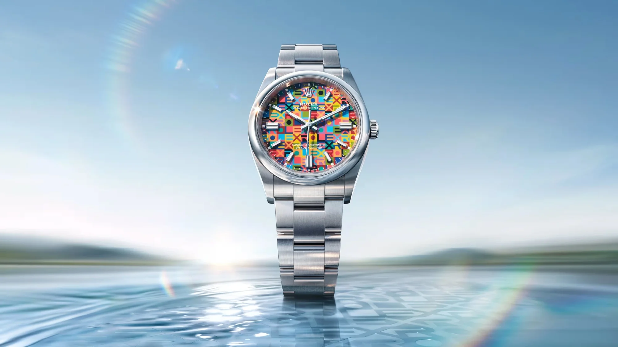 Rolex Oyster Perpetual 36 modelinin parlak çelik kasası ve bileziği ile öne çıkan, canlı mavi, sarı, turuncu, yeşil, pembe ve kırmızı tonlardaki geometrik desenler ve tekrar eden Rolex logolarıyla bezenmiş renkli kadranı, hafif dalgalı su yüzeyi ve açık mavi gökyüzü fonunda etkileyici bir şekilde sergileniyor.