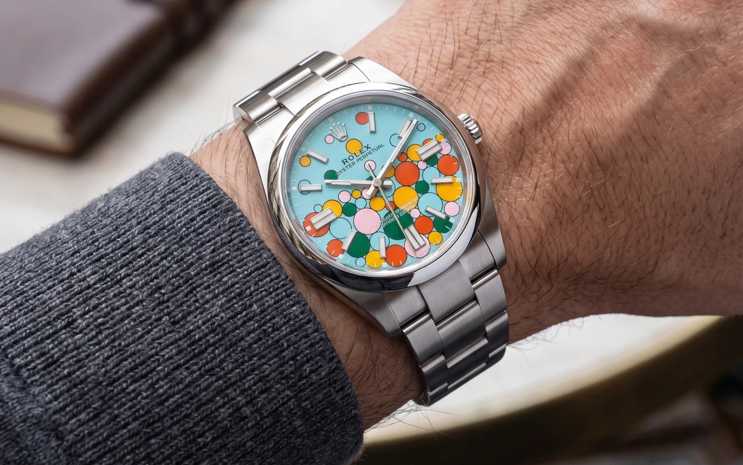 Açık mavi zemin üzerinde pembe, sarı, turuncu ve yeşil renklerdeki balon benzeri desenlerle bezeli Rolex Oyster Perpetual Celebration kadran saati, gri örgülü bir kazağın kolundan görünen bir bilekte şıkça duruyor.