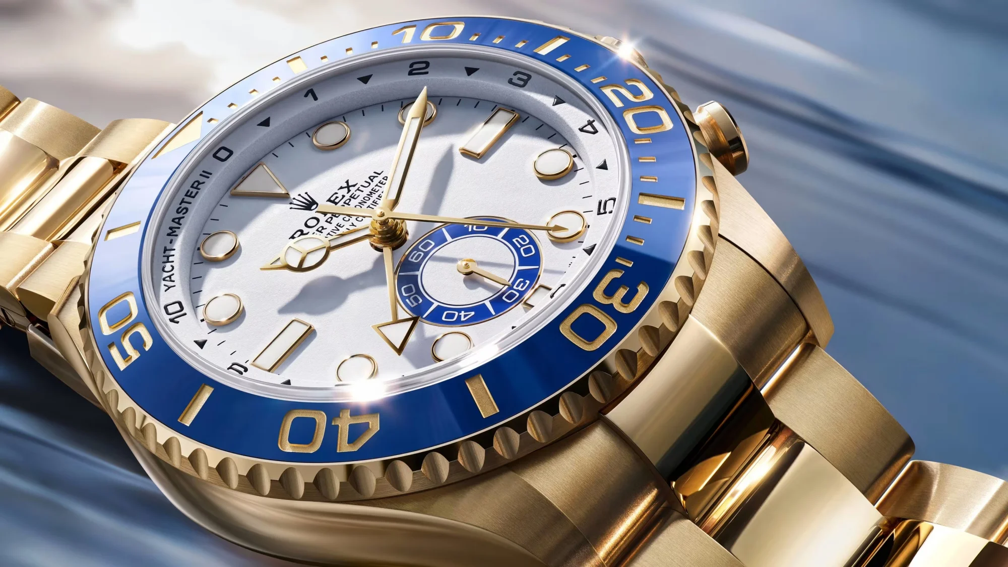 Watches and Wonders etkinliğinden, parlak sarı altın kasası ve bileziği olan, beyaz kadranında Rolex Yacht-Master II markası belirginleşen, mavi seramik bezelindeki altın rakamlar ve alt kadranıyla dikkat çeken, dinamik mavi bir arka plan üzerinde zarif bir saat yakın çekim sunuyor.