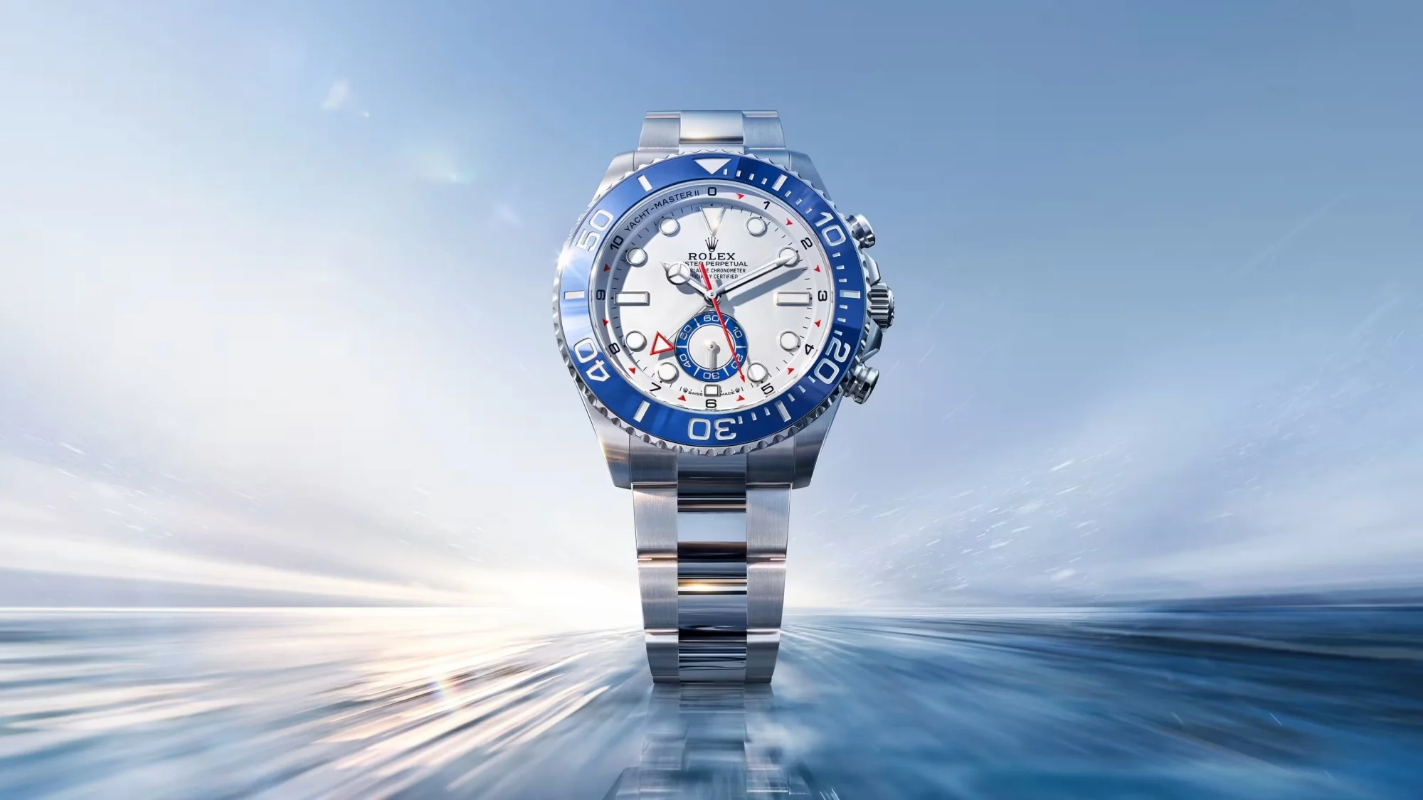 Parlak, gümüş renkli paslanmaz çelik kasa ve bileziğe sahip bir Rolex Yacht-Master II saati, beyaz kadranı ve dikkat çekici mavi regatta kronograf bezeliyle dinamik, bulanık açık mavi bir arka plan önünde hız ve denizcilik temasını vurgulayarak şık bir şekilde sergileniyor.