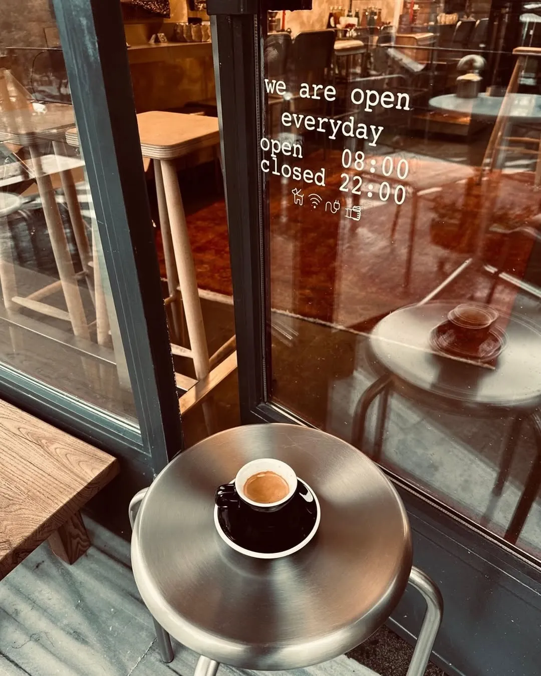 İçerideki modern kafe ortamının yansıdığı cam pencerenin önünde, üzerinde günlük 08:00-22:00 çalışma saatleri yazan bir metal taburede, sıcak ve koyu renkli bir fincan espresso misafirleri taze bir kahve molasına davet ediyor.