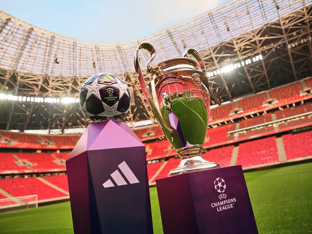 Boş bir futbol stadyumunun yemyeşil sahasında, Adidas logolu mor bir podyumda duran UEFA Şampiyonlar Ligi futbol topu ve onun yanında Şampiyonlar Ligi kupası, yaklaşan büyük finalin heyecanını ve festival atmosferini çağrıştırıyor.