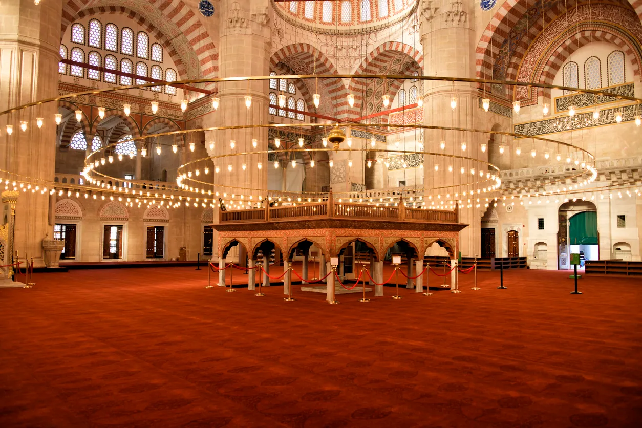 Selimiye Camii'nin geniş ve görkemli iç mekanından bir kare; kırmızı halılarla döşeli zeminin ortasında, detaylı ahşap işçiliğine sahip müezzin mahfili tüm zarafetiyle yükselirken, tavanlardan sarkan yüzlerce ampullü dairesel avizeler mekanı sıcak bir ışıltıyla aydınlatıyor.