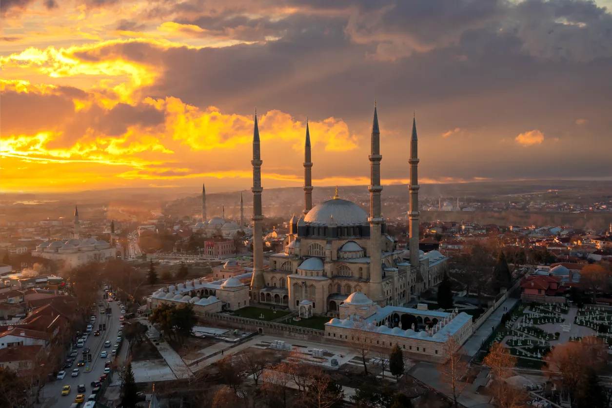 Selimiye Camii, altın sarısı ve turuncu bulutlarla kaplı dramatik bir gün batımı veya doğumu manzarasının önünde, dört ince minaresi ve büyük kubbesiyle çevresindeki şehir yapılarının ve hareketli yolların üzerinde görkemli bir şekilde yükseliyor.