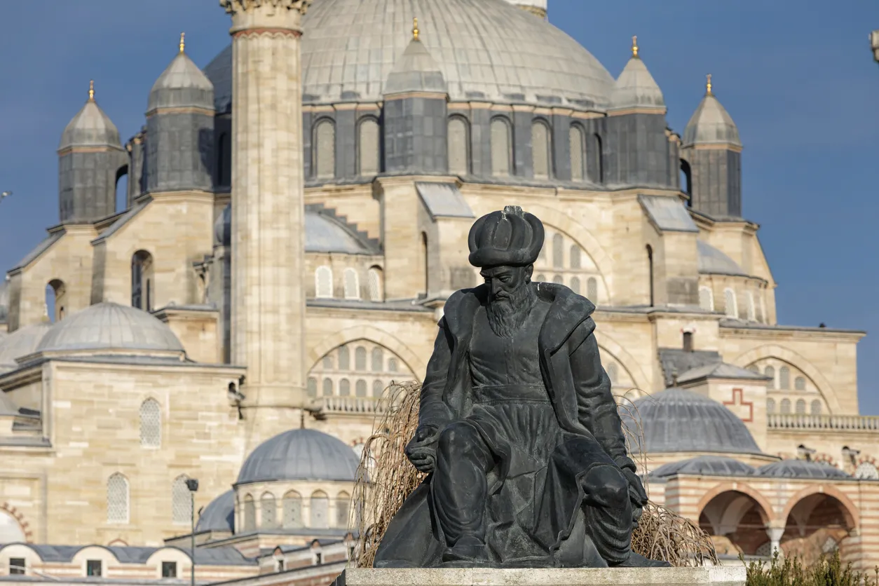 Mimar Sinan'ın sakallı, sarıklı ve kaftanlı koyu renkli heykeli ön planda oturur pozisyonda dururken, arka planda güneşli mavi gökyüzü altında Edirne'deki Selimiye Camii'nin görkemli ana kubbesi, yan kubbeleri ve bir minaresi belirgin bir şekilde yükseliyor.