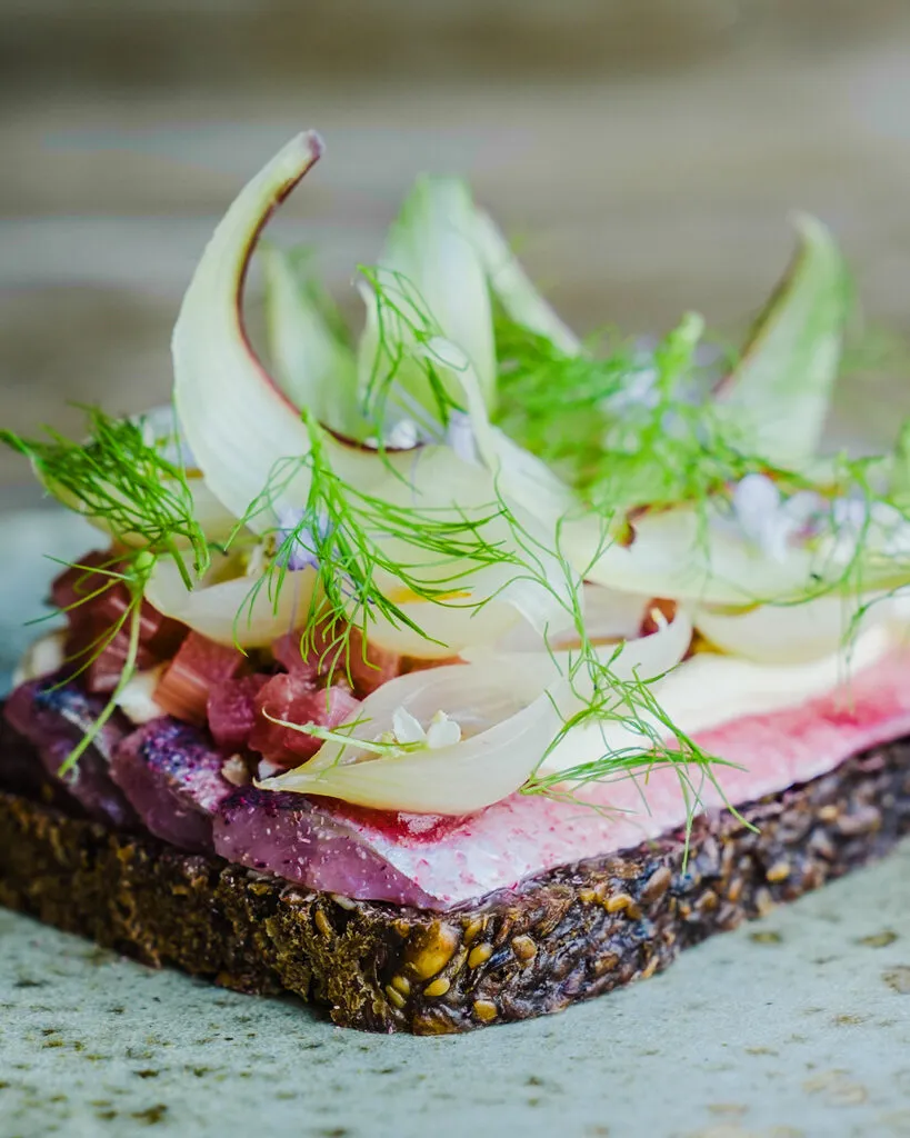 Selma restoranından enfes bir örnek olan bu smørrebrød, koyu renkli, tanecikli çavdar ekmeği üzerinde pembe tonlarda marine edilmiş ringa balığı dilimleri, kremsi beyaz sos, zarifçe kıvrılmış rezene dilimleri ve bolca taze dereotuyla süslenmiş, küçük mor çiçeklerle detaylandırılmış eşsiz bir Kopenhag lezzetini gözler önüne seriyor.
