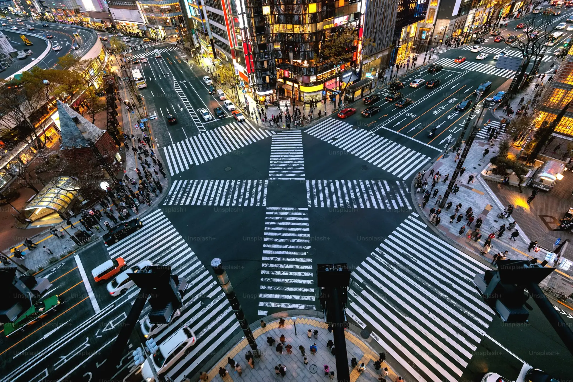 Akşamın alacakaranlığında, Tokyo'nun ikonik Shibuya geçidindeki yüksek açılı bu görünümde, kalabalık yaya trafiği ve araçlar, etrafı neon ışıklı tabelalar ve çok katlı binalarla çevrili karmaşık zebra geçitleri üzerinde dinamik bir şehir hareketliliği oluşturuyor.