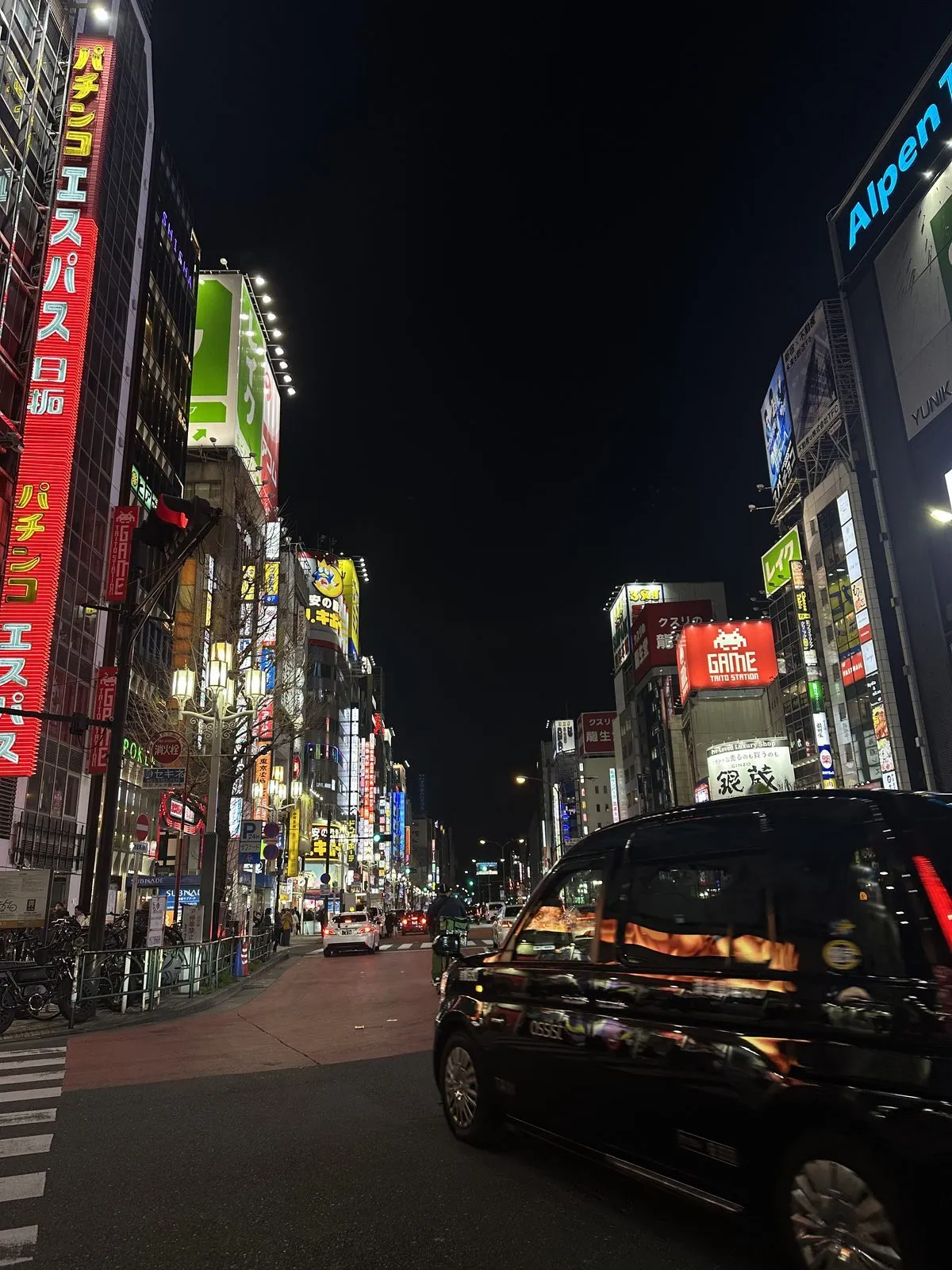 Shibuya gecesinde çekilmiş bu geniş açılı fotoğraf, neon ışıklı Japonca tabelalarla bezeli yüksek binaların arasından ilerleyen hareketli bir caddeyi gösteriyor, sağ önde geçen siyah bir araç şehrin dinamik ve canlı atmosferini pekiştiriyor.