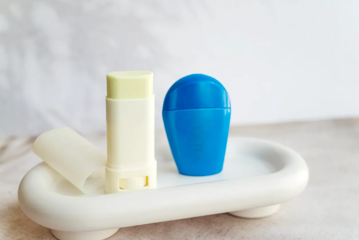Beyaz, minimalist bir tepsi üzerinde, kapağı açık, hafif yeşilimsi sarı renkli ucu görünen bir stick güneş koruyucu ile yanında duran parlak mavi, kubbe şekilli bir başka stick güneş koruyucu, ürünlerin taşınabilir ve pratik yapısını vurguluyor.