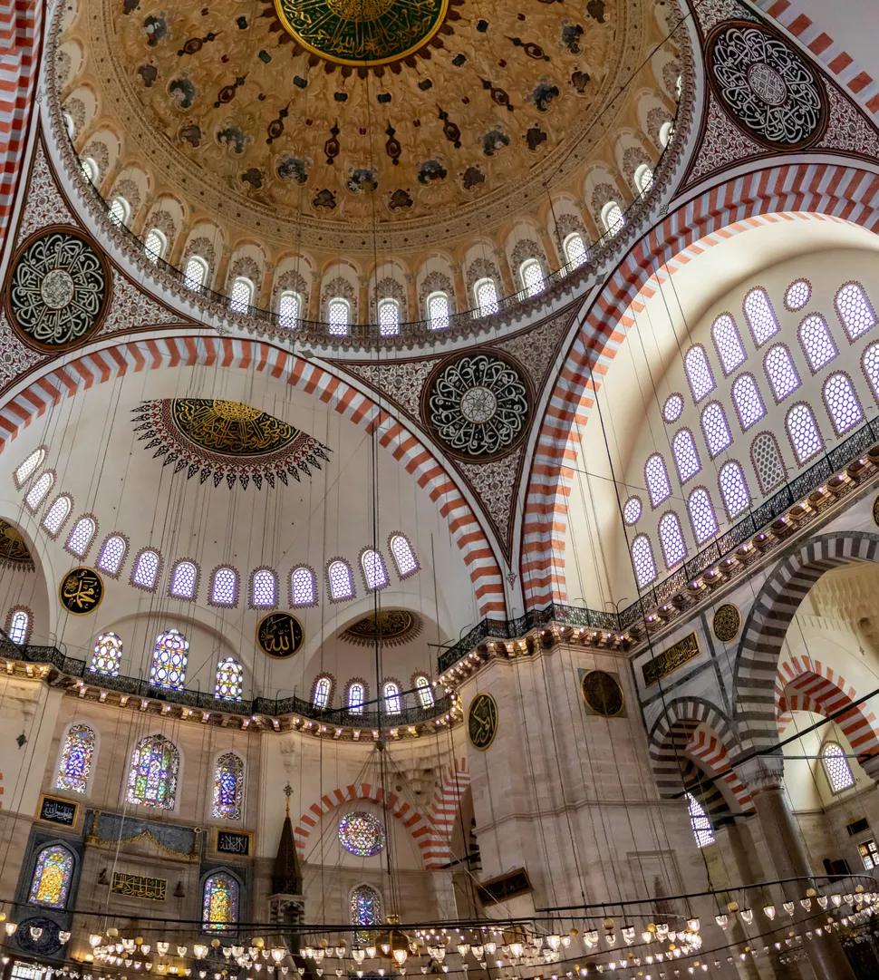 Süleymaniye Camii'nin büyüleyici iç mekanından, altın rengi ve yeşil hat sanatı işlemeleriyle bezeli muhteşem ana kubbesi, kubbeyi çevreleyen sıralı kemerli pencereler, kırmızı beyaz çizgili destek kemerleri, alt katlardaki renkli vitraylar ve zarif sarkıt avizelerin görkemli bir panoraması sergileniyor.