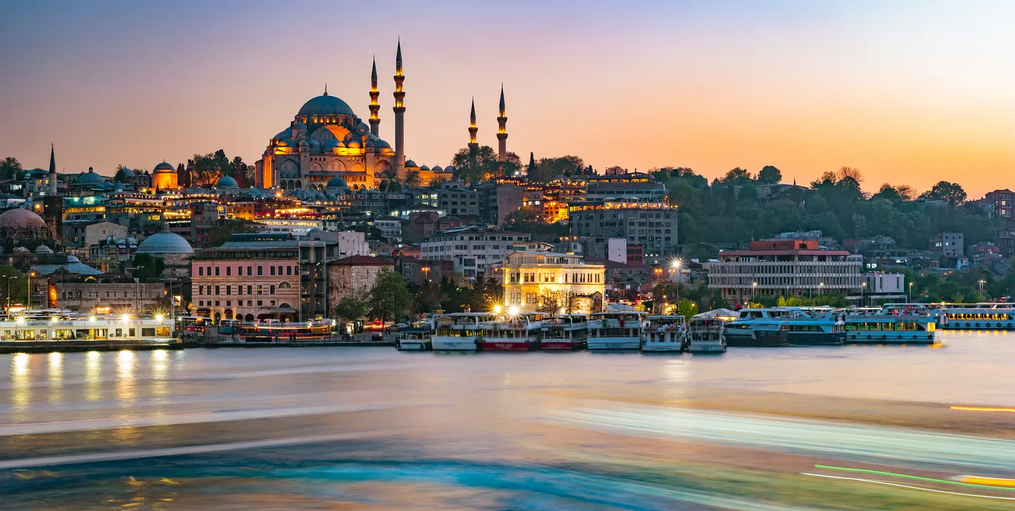Gün batımında İstanbul siluetini gözler önüne seren bu geniş açılı çekimde, ışıklandırılmış Süleymaniye Camii'nin heybetli kubbe ve minareleri şehrin tarihi dokusuyla uyum içinde yükselirken, Boğaz'ın hareketli sularında teknelerin oluşturduğu uzun ışık izleri ve yansımaları büyüleyici bir atmosfer sunuyor.