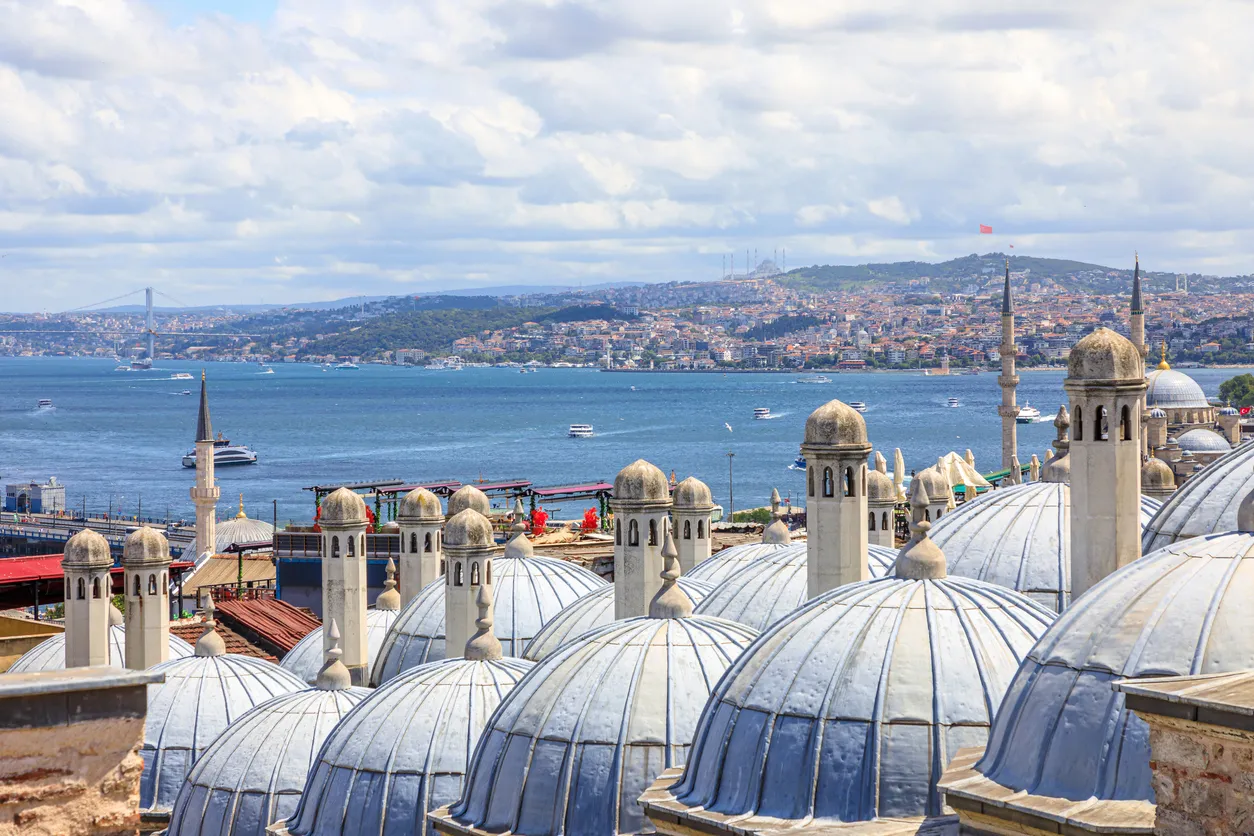 Süleymaniye Camii'nin ön plandaki kurşuni kubbeleri ve yükselen minareleri arasından görünen muhteşem İstanbul Boğazı manzarası; Boğaz'da seyreden çok sayıda gemi, ilerideki köprü ve bulutlu gökyüzü altında uzanan şehrin panoramik görüntüsünü sunuyor.