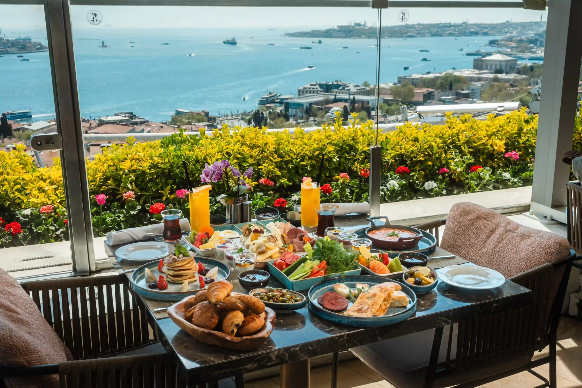 Çeşit çeşit lezzetlerle donatılmış zengin bir kahvaltı sofrası, Boğaz'ın ve üzerinde gezen teknelerin nefes kesen panoramik İstanbul manzarasına nazır, rengarenk çiçeklerle çevrili bir teraslı mekânda güne keyifli bir başlangıç yapmak için misafirlerini bekliyor.