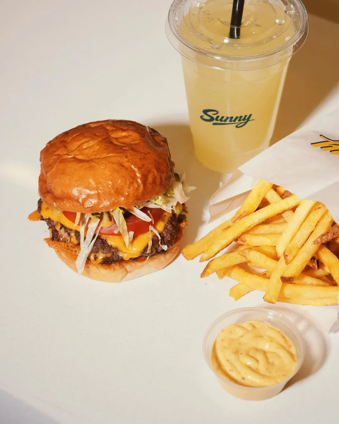 Beyaz bir zeminde, erimiş peynirli, domatesli ve marullu iki köfteli, parlak ekmekli lezzetli bir smash burger, bir kesekağıdından taşan altın sarısı çıtır patates kızartmaları, küçük bir kapta kremsi sarı bir sos ve siyah pipetli şeffaf bardakta, üzerinde Sunny markası yazan ferahlatıcı limonata iştah açıcı bir şekilde duruyor.