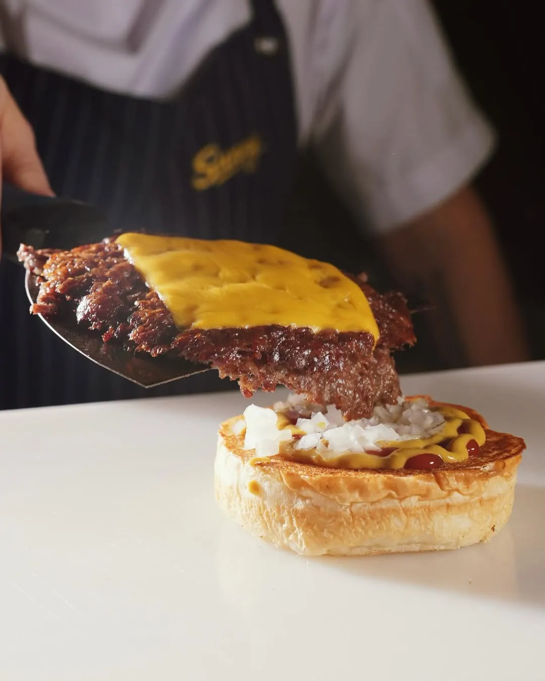 Siyah önlüğünde Sunny logosu bulunan bir şefin metal bir spatula yardımıyla erimiş sarı peynirle kaplı, ince ve kızarmış bir smash burger köftesini, bolca doğranmış soğan, sarı hardal ve kırmızı ketçap sürülmüş kızarmış hamburger ekmeğinin alt katmanına dikkatlice yerleştirdiği bir an gösteriliyor.
