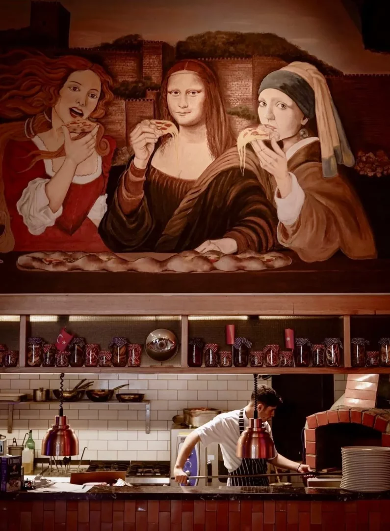 Suzgeç Marina mutfağının üzerinde, Venüs, Mona Lisa ve İnci Küpeli Kız'ın pizza yerken tasvir edildiği büyük, esprili bir duvar resmi yer alırken; alt kısımda bir şef geleneksel tuğla fırının başında özenle yemek hazırlıyor ve raflarda kavanozlar sıralanıyor.