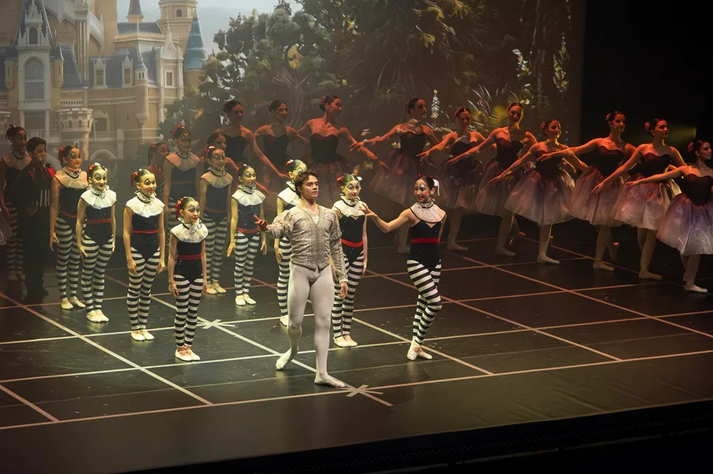 Kuğu Gölü balesi performansı sırasında, arka planda şato ve orman dekoruyla aydınlatılmış sahnede, fildişi renkli kostümlü erkek dansçı ile siyah beyaz çizgili giysili ve fırfırlı yakalı kadın dansçı liderliğindeki çocuklar ve tütülü balerinler uyumlu bir gösteri sunuyor.