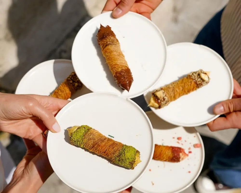 Gastronomi buluşması Taste and the City'de, fıstıklı, çikolatalı ve farklı dolgularla zenginleştirilmiş beş lezzetli kadayıf rulosunun tadım ve paylaşım anını yansıtan, katılımcıların ellerinde tuttuğu tabakların yakın çekimi.