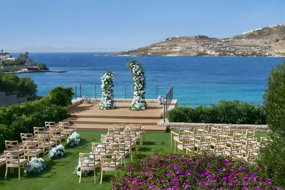 The Bodrum Edition'ın deniz manzaralı yemyeşil bir terasında, beyaz çiçeklerle süslenmiş seramoni alanı ve davetli sandalyeleri ile kusursuz bir düğün deneyimi için düzenlenmiş, Ege'nin turkuaz sularına bakan pastoral bir açık hava düğün mekânı.