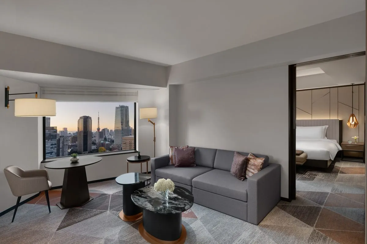 Modern bir Tokyo otel süitinin ferah oturma alanı, pencereden görünen ikonik Tokyo silüeti ve Tokyo Kulesi manzarası eşliğinde konforlu gri bir kanepe, şık sehpa ve yemek masasıyla lüks bir yaşam hissi sunuyor, arka planda ise yatak odası zarifçe beliriyor.