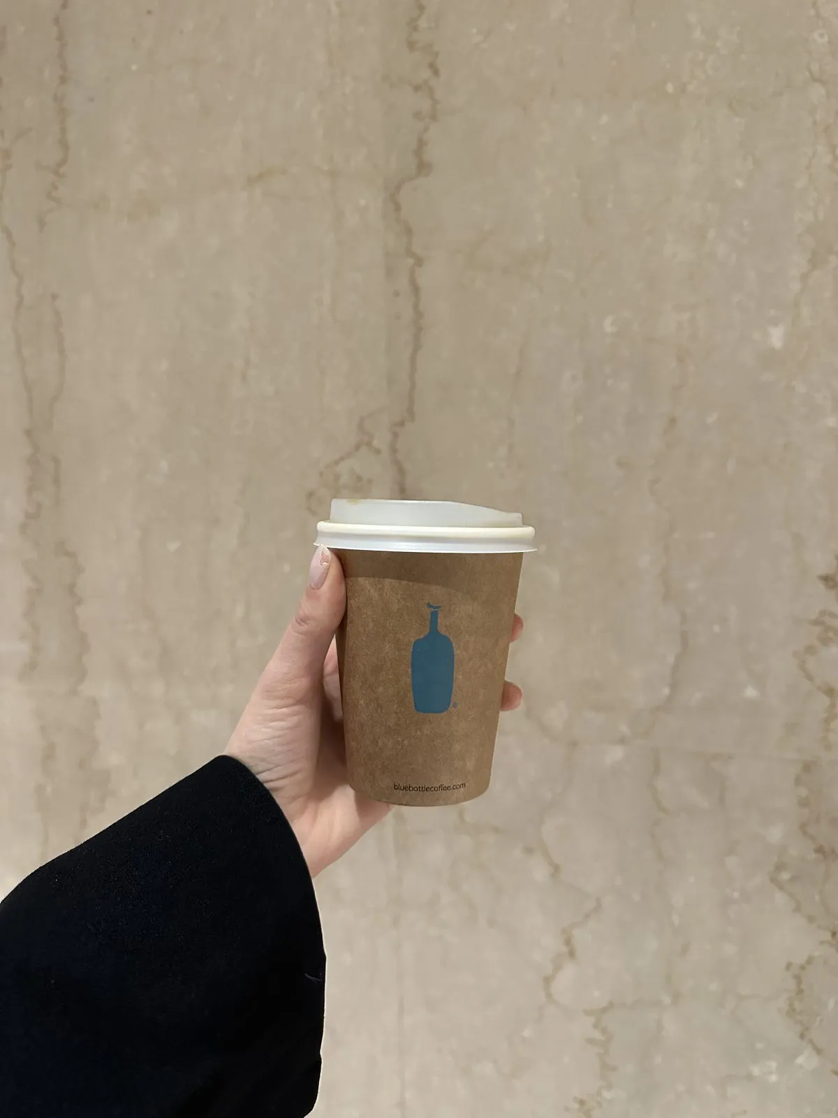 Açık bej mermer dokulu bir duvarın önünde, siyah bir kıyafet giyen bir el, üzerinde mavi şişe logosu ve bluebottlecoffee.com adresi bulunan, beyaz kapaklı kahverengi bir Blue Bottle kahve bardağını nazikçe tutuyor ve bu kare, Tokyo seyahatinde keşfedilen nitelikli kahvecilerdeki keyifli bir kahve molasını gözler önüne seriyor.