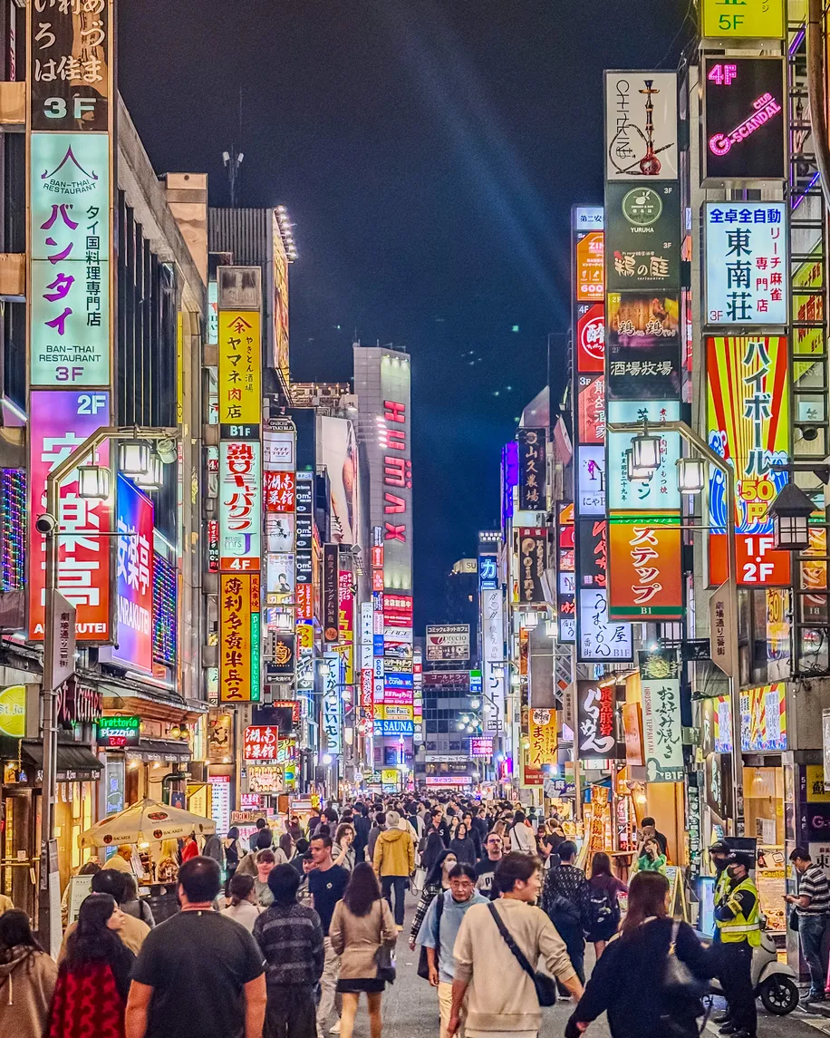 Tokyo seyahati sırasında görülebilecek, geceleri rengarenk neon ışıkları ve devasa reklam panolarıyla göz kamaştıran, hareketli bir caddede yürüyen kalabalıklar, şehrin eşsiz ve enerjik atmosferini canlı bir şekilde yansıtıyor.