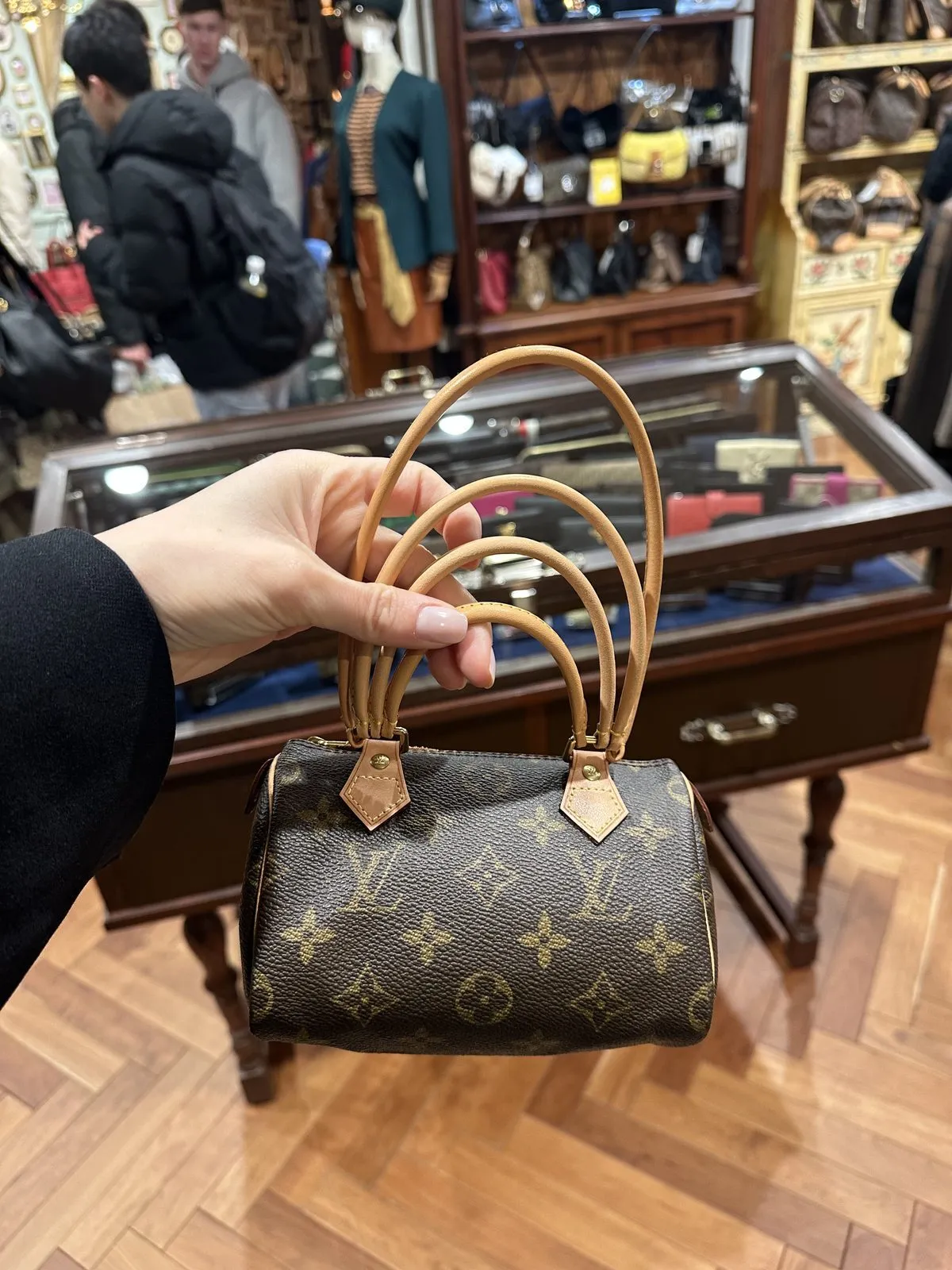 Bir el, klasik monogram desenli, minyatür kahverengi bir Louis Vuitton çantayı açık renk deri saplarından tutuyor ve arka planda Tokyo'daki bir vintage mağazasının bulanık görüntüsüyle, ahşap vitrinler ve sergilenen diğer çantalar seçilebiliyor.
