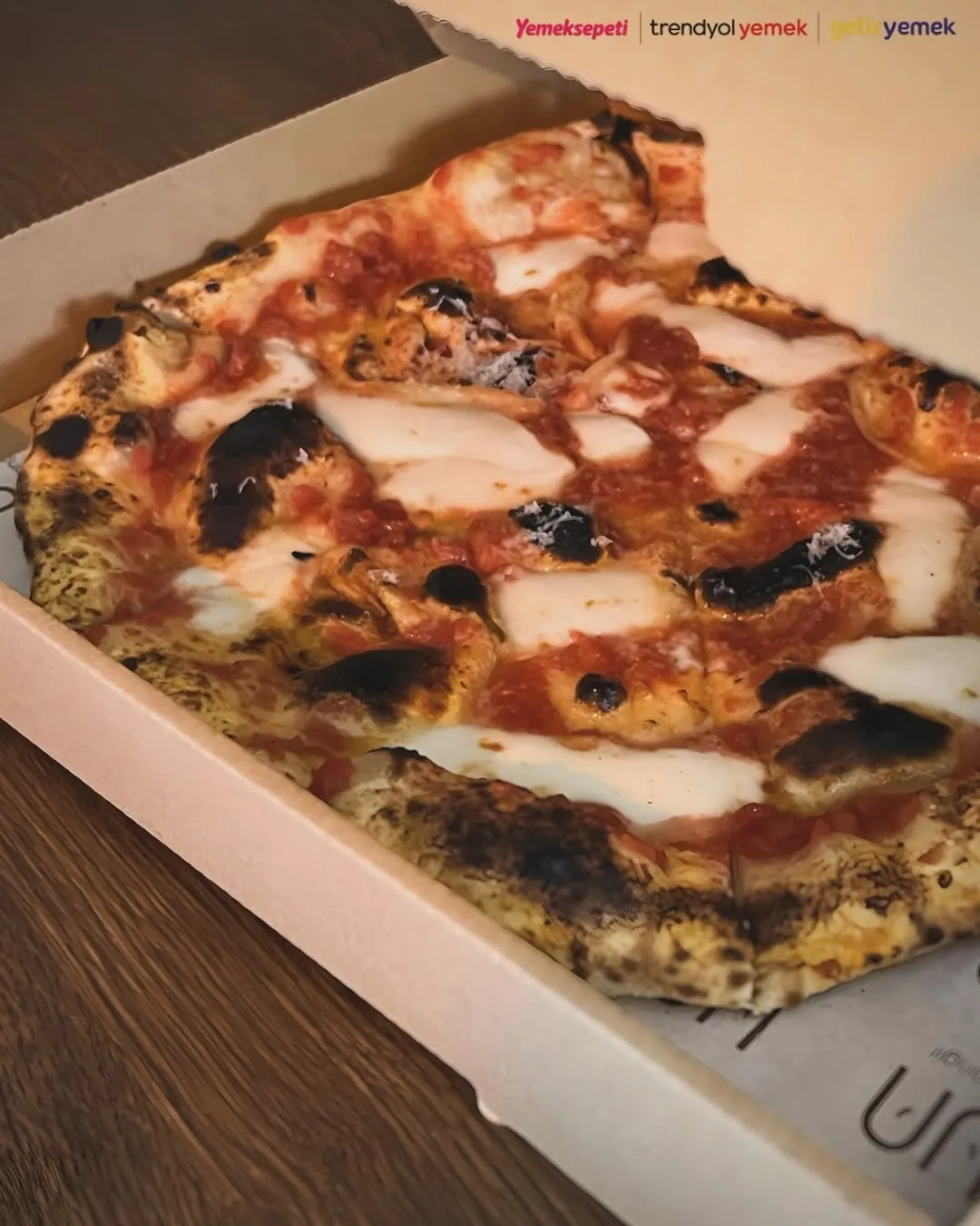 Cihangir'deki UN restoranından Yemeksepeti logosu görünen bir kutu içerisinde servis edilen, kenarları odun ateşinde pişmiş gibi hafifçe kömürleşmiş, bol domates sosu ve erimiş mozzarella peyniriyle bezeli, sıcak ve iştah açıcı bir pizzanın yakın çekimi.