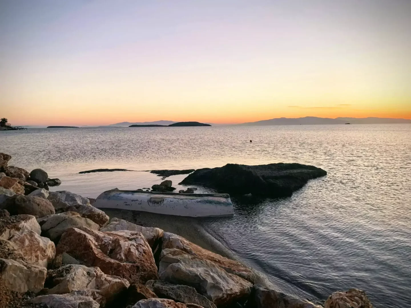Urla'da gün batımının huzurlu atmosferinde, turuncu ve mor tonlarıyla parlayan gökyüzünün altında, kıyıya vurmuş devrilmiş beyaz bir tekne ve kayalıklar yer alırken, dingin deniz ve uzaklardaki adaların silüetleri göz alıcı bir manzara oluşturuyor.