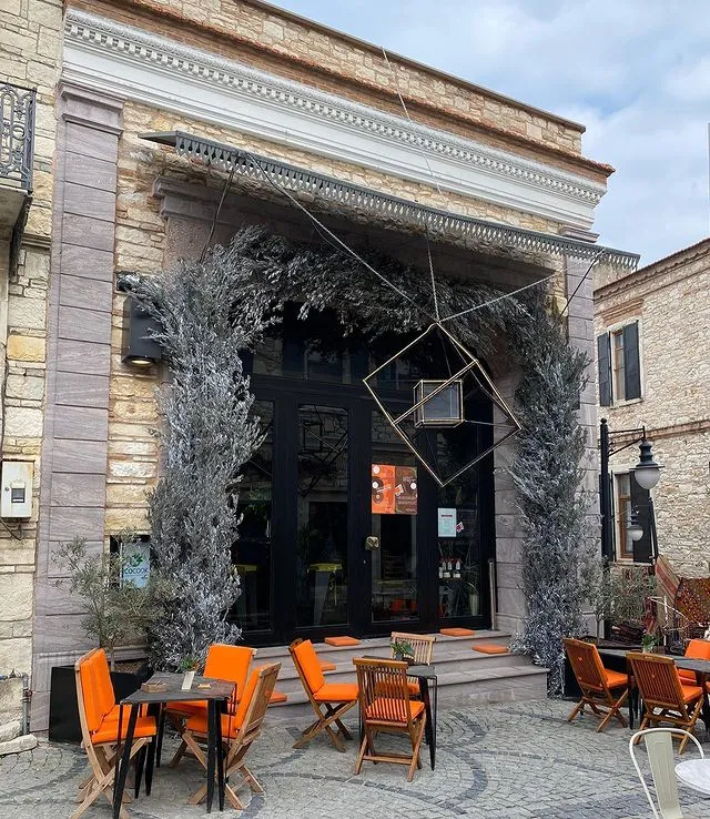 Urla'daki taş cepheli, gümüşi yapraklı ağaçlarla çevrili şık bir mekanın önünde, modern geometrik bir metal heykelin asılı olduğu siyah kapılı girişi ve kaldırımda parlak turuncu minderli ahşap sandalyelerle dolu davetkar açık hava oturma alanı, bu lezzetli mekanın kendine özgü atmosferini sergiliyor.