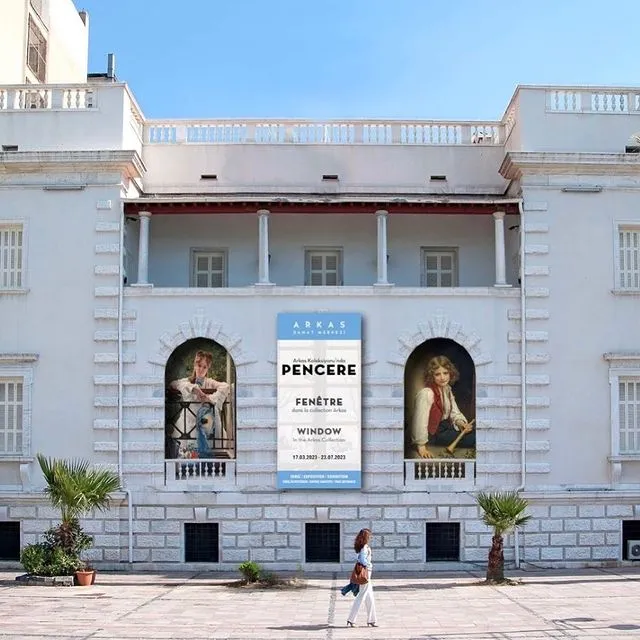 Güneşli bir havada, beyaz klasik mimarili Arkas Sanat Merkezi binasının önünde yürüyen bir kadın ve merkezin PENCERE adlı sergisini duyuran büyük afişiyle birlikte, kemerli pencerelerde sergilenen tablolar dikkat çekiyor.