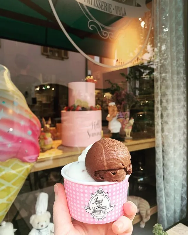 Urla'daki lezzetli bir pastaneden alınan, pembe puantiyeli Irmik Hanım PATISSERIE - URLA logolu kapta çikolatalı ve vanilyalı dondurma, arka planda vitrindeki Hello Summer yazılı pembe pasta ve sevimli figürlerle birlikte keyifli bir anı yansıtıyor.
