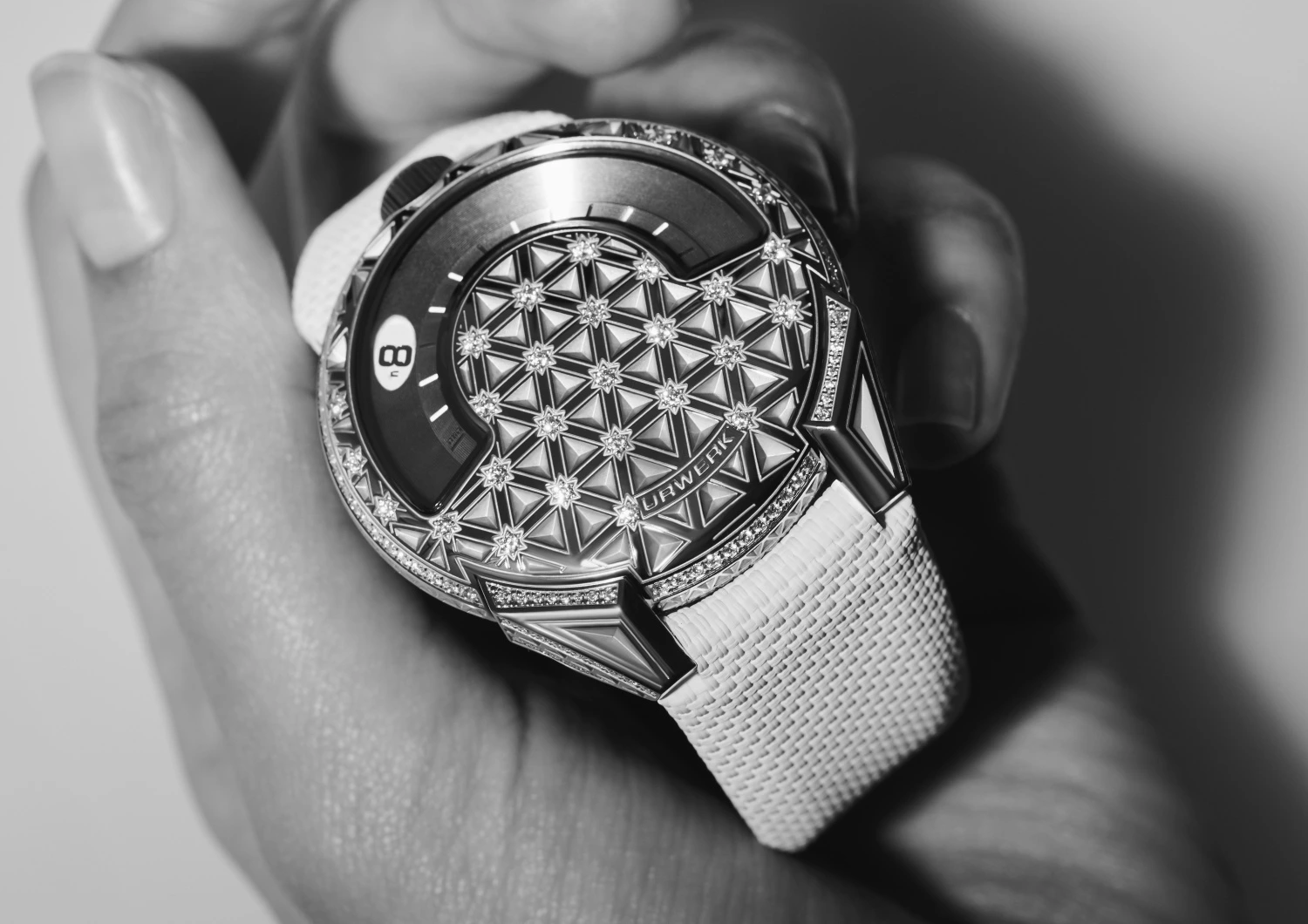 Siyah beyaz fotoğrafta, bir el lüks URWERK UR-101 saati tutuyor; saatin kadranı ve çerçevesi, geometrik bir desen oluşturan çok sayıda parlak pırlanta ile süslenmiş, benzersiz tasarımı ve saat 8'i gösteren dijital ekranı dikkat çekiyor.
