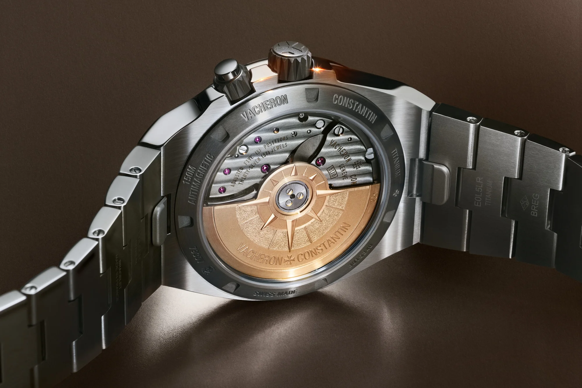 Parlak metal kordonlu Vacheron Constantin Overseas Dual Time saatin arkadan görünümünde, şeffaf arka kapağın ardında, kardinal yönleri gösteren altın renkli rotoru ve hassas iç mekanizmasının detaylı işçiliği tüm ihtişamıyla sergileniyor.