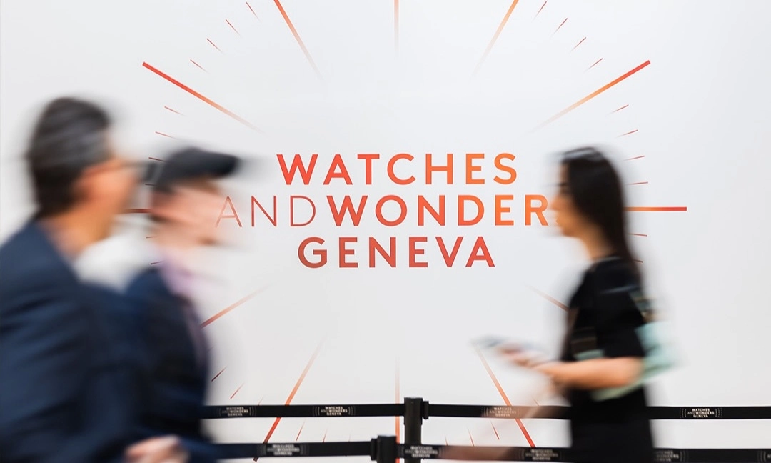 Büyük beyaz bir duvarda, ortasında turuncu renkte WATCHES AND WONDERS GENEVA yazısı ve etrafında yayılan saat ibrelerini andıran çizgiler bulunurken, önünden geçen bulanıklaşmış ziyaretçi silüetleri Watches and Wonders etkinliğinin hareketliliğini yansıtıyor.