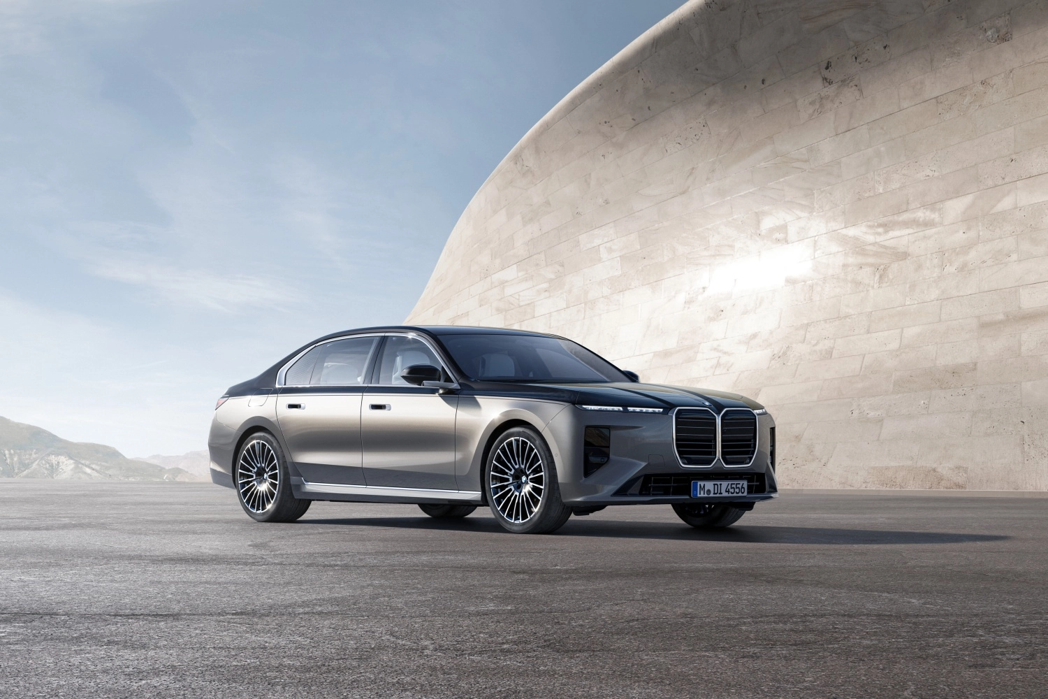 Yeni BMW 7 Serisi'nin şık ve dikkat çekici çift renkli gövdesi, modern mimarinin ve uzaktaki dağların eşlik ettiği geniş bir dış mekanda ön planda duruyor, far detayları ve geniş ızgarasıyla lüks bir görünüm sunuyor.