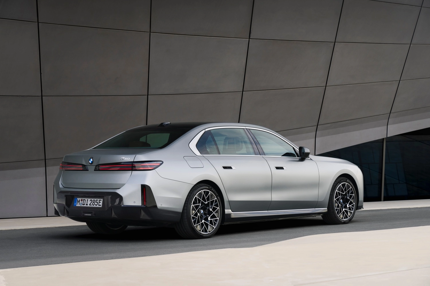 Parlak gri renkteki, çift renkli (gri ve siyah tavan) yeni BMW 7 Serisi otomobilin modern mimariye sahip bir binanın önünde dururken çekilmiş, belirgin ince arka stop lambaları ve çarpıcı desenli jantlarıyla arka üç çeyrek tasarımı.