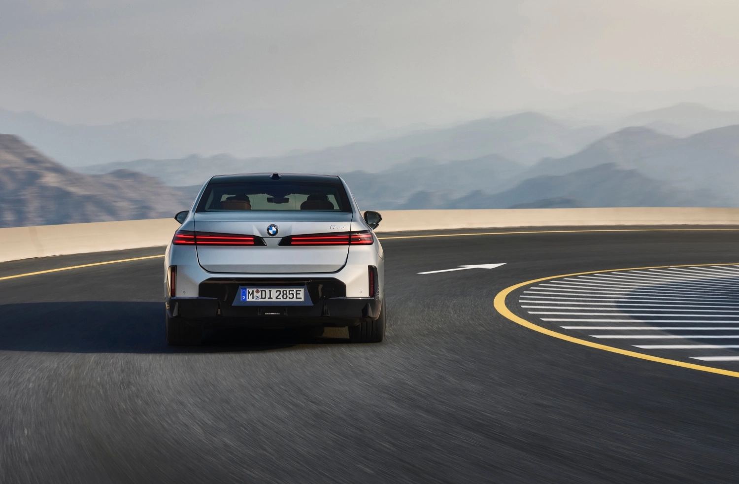 Gümüş rengi yeni BMW 7 Serisi i7, modern ve belirgin yatay arka lambalarıyla dikkat çeken dış tasarımıyla, kıvrımlı asfalt bir yolda ilerlerken arka kısmından çekilmiş bir fotoğrafta, sisli dağ manzarası eşliğinde dinamik bir sürüş anını yansıtıyor.