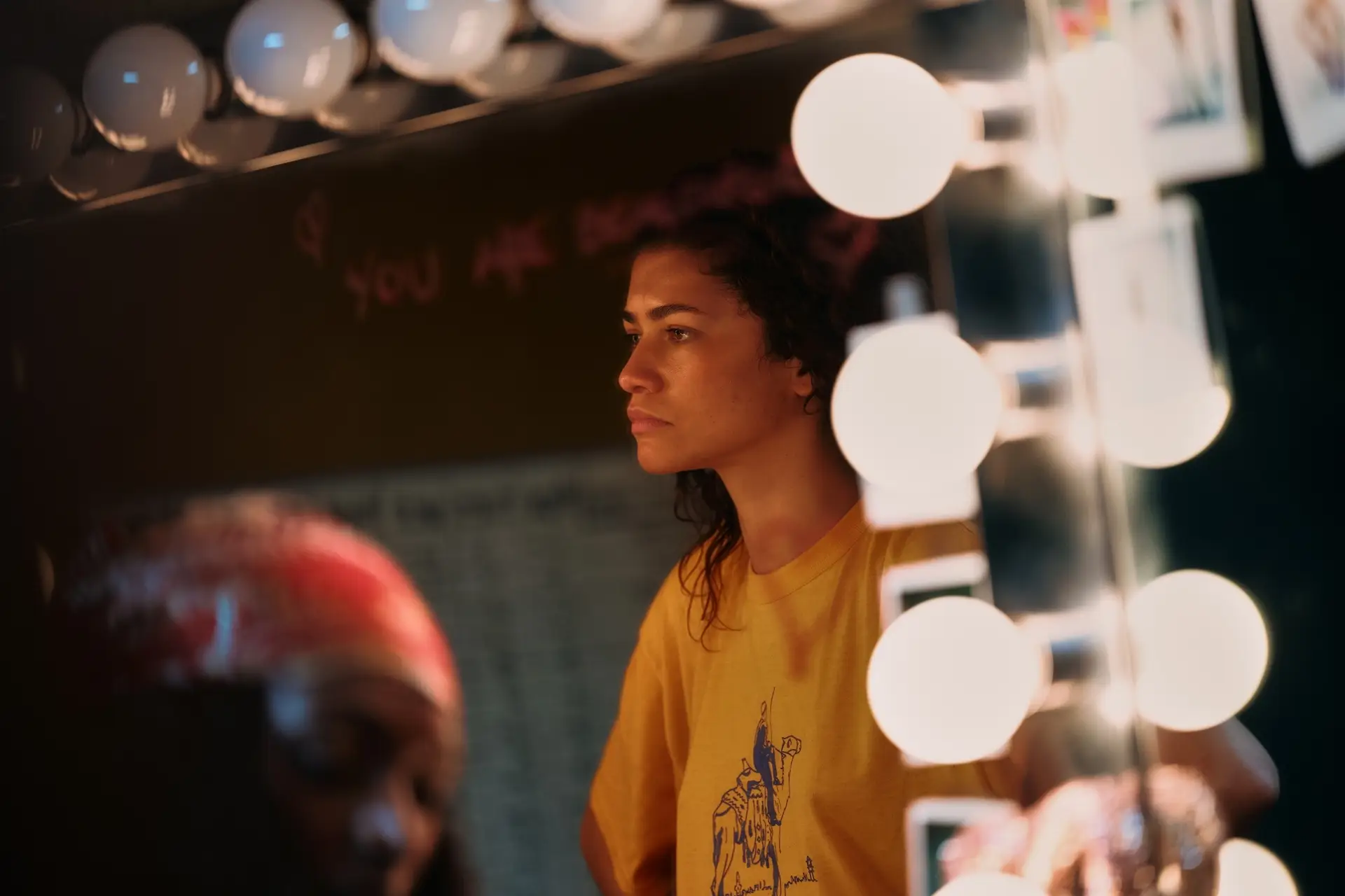 Karanlık bir odada, makyaj ışıklarının çevrelediği bir aynanın önünde sağ profilden görünen Zendaya, Euphoria dizisindeki Rue karakteri olarak düşünceli bir ifadeyle ileriye bakıyor, sarı tişörtü ve dağınık kıvırcık saçlarıyla yüzünde sıcak aydınlık parlamalar var.