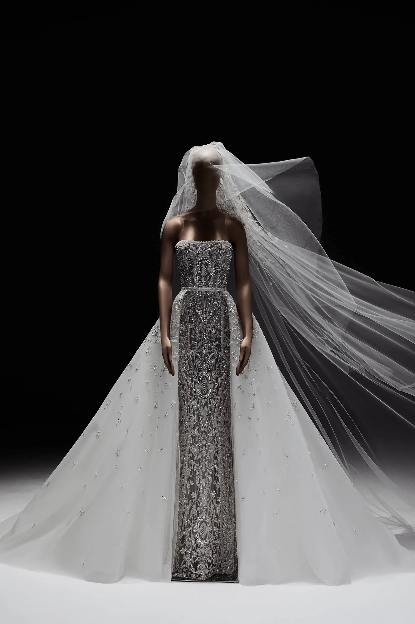 New York Bridal Fashion Week'in öne çıkan yeni sezon gelinlik trendlerinden biri olarak, bir manken üzerinde sunulan bu Elie Saab 2027 İlkbahar gelinliği, gümüş işlemeli straplez dar kesim iç elbiseyi saran hacimli, pırıltılı beyaz tül dış eteği ve savrulan uzun duvağıyla masalsı bir zarafet sergiliyor.