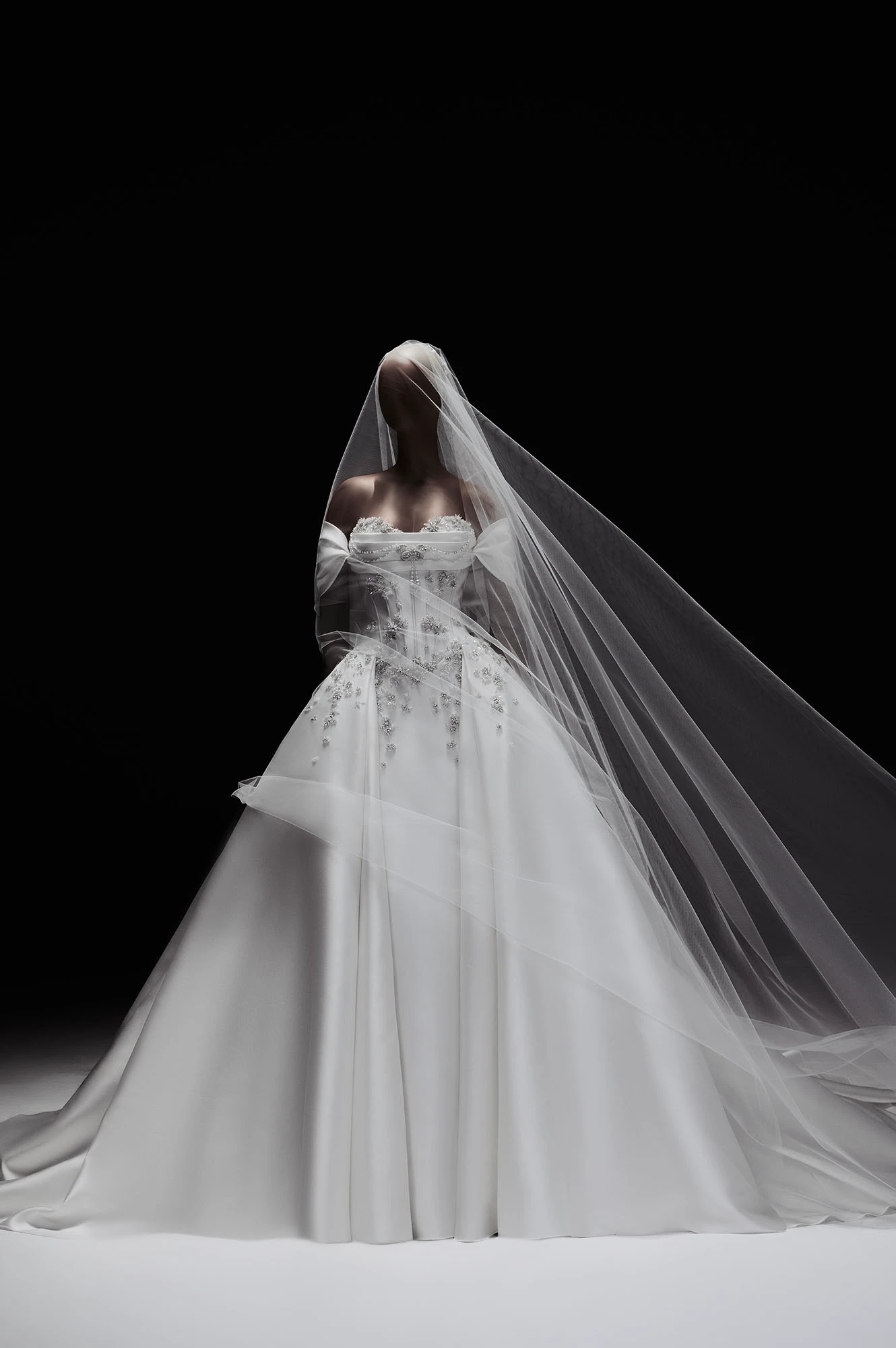Elie Saab 2027 İlkbahar gelin koleksiyonundan, zarif nakışlarla bezeli korseli bir üst kısma, volümlü eteklere ve rüzgarda süzülen uzun şeffaf bir duvağa sahip, karanlık fonda adeta yaşayan bir kompozisyon gibi duran görkemli bir balo gelinliği.