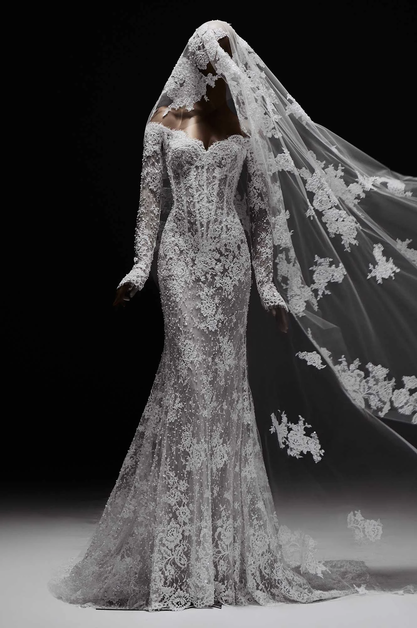 Elie Saab'ın 2027 İlkbahar gelinlik koleksiyonundan, omuzları açık, uzun kollu, vücudu saran yoğun dantel işlemeler ve ışıltılı boncuklarla süslü mermaid kesim bir gelinlik, rüzgarda dalgalanan uzun, dantel detaylı duvağıyla büyüleyici ve zamansız bir bridal şıklık sunuyor.