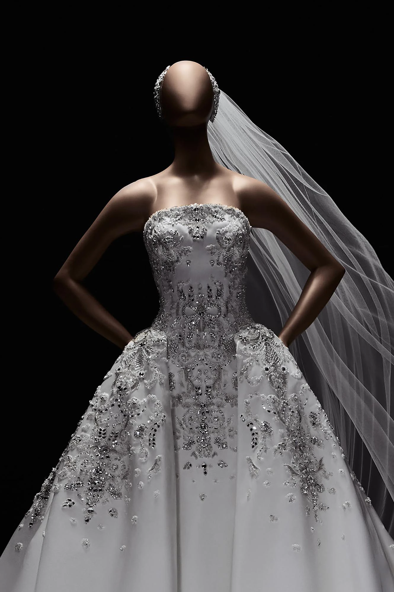 Başsız, bronz tenli bir manken üzerinde, gümüş işlemeli ve taş detaylı, straplez, prenses kesim beyaz bir Elie Saab 2027 İlkbahar gelinlik modeli görülüyor; bu göz alıcı tasarım, zarif dantel ve boncuk işlemeleriyle 2027 gelinlik trendlerini yansıtırken, mankenin belinde elleri ve arkaya doğru hafifçe uçuşan tül duvağı dramatik siyah fonda etkileyici bir estetik sunuyor.