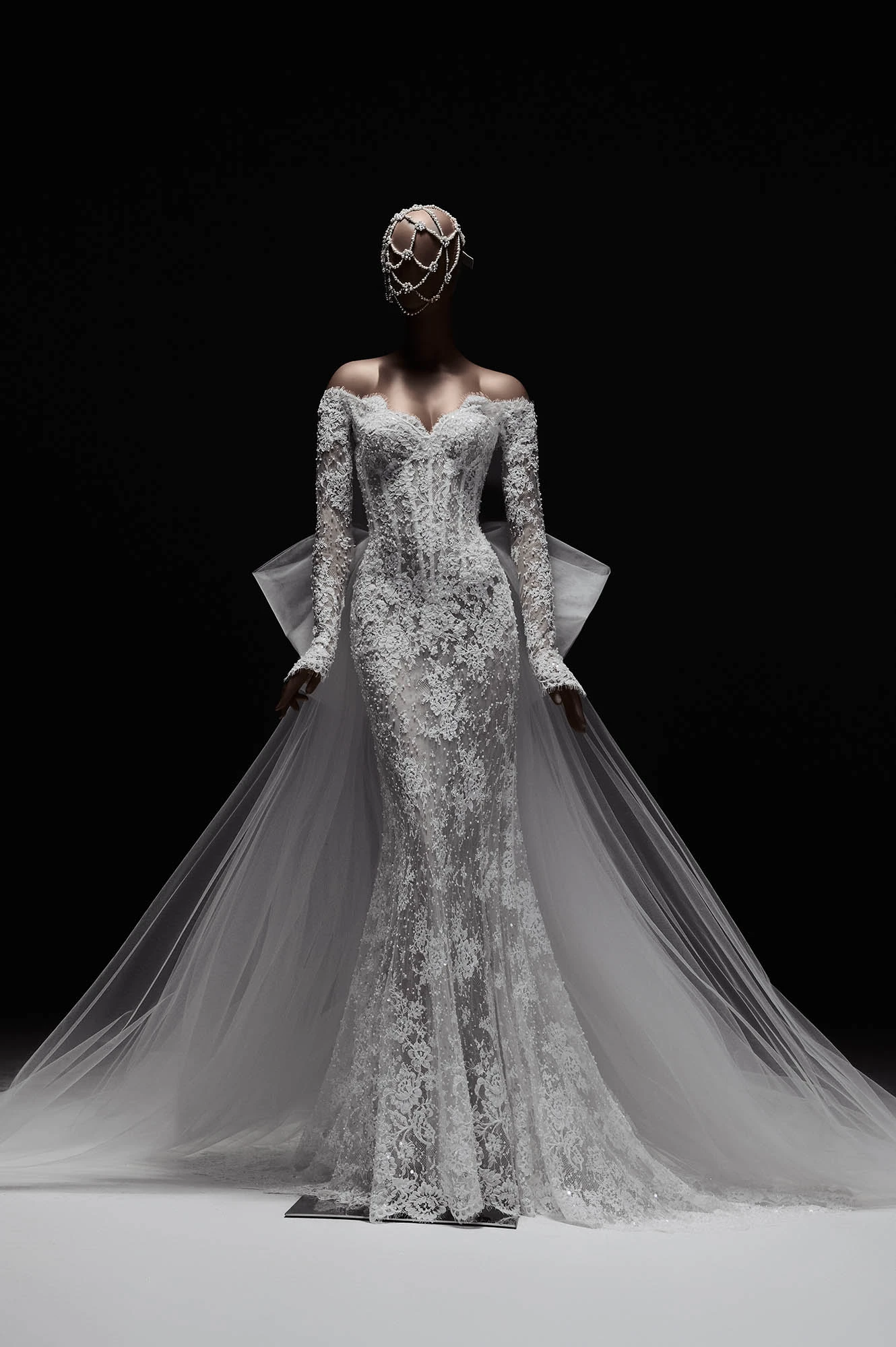 Elie Saab 2027 İlkbahar Gelinlik Koleksiyonu'ndan, omuzları açık, uzun kollu, baştan aşağı narin dantel motifleri ve ışıltılı inci detaylarıyla süslenmiş, vücudu saran, arkasında hacimli bir fiyonk ve uzun tül kuyrukla tamamlanan, inci işlemeli ağ tarzı bir başlık taşıyan manken üzerindeki masalsı görkemli gelinlik.
