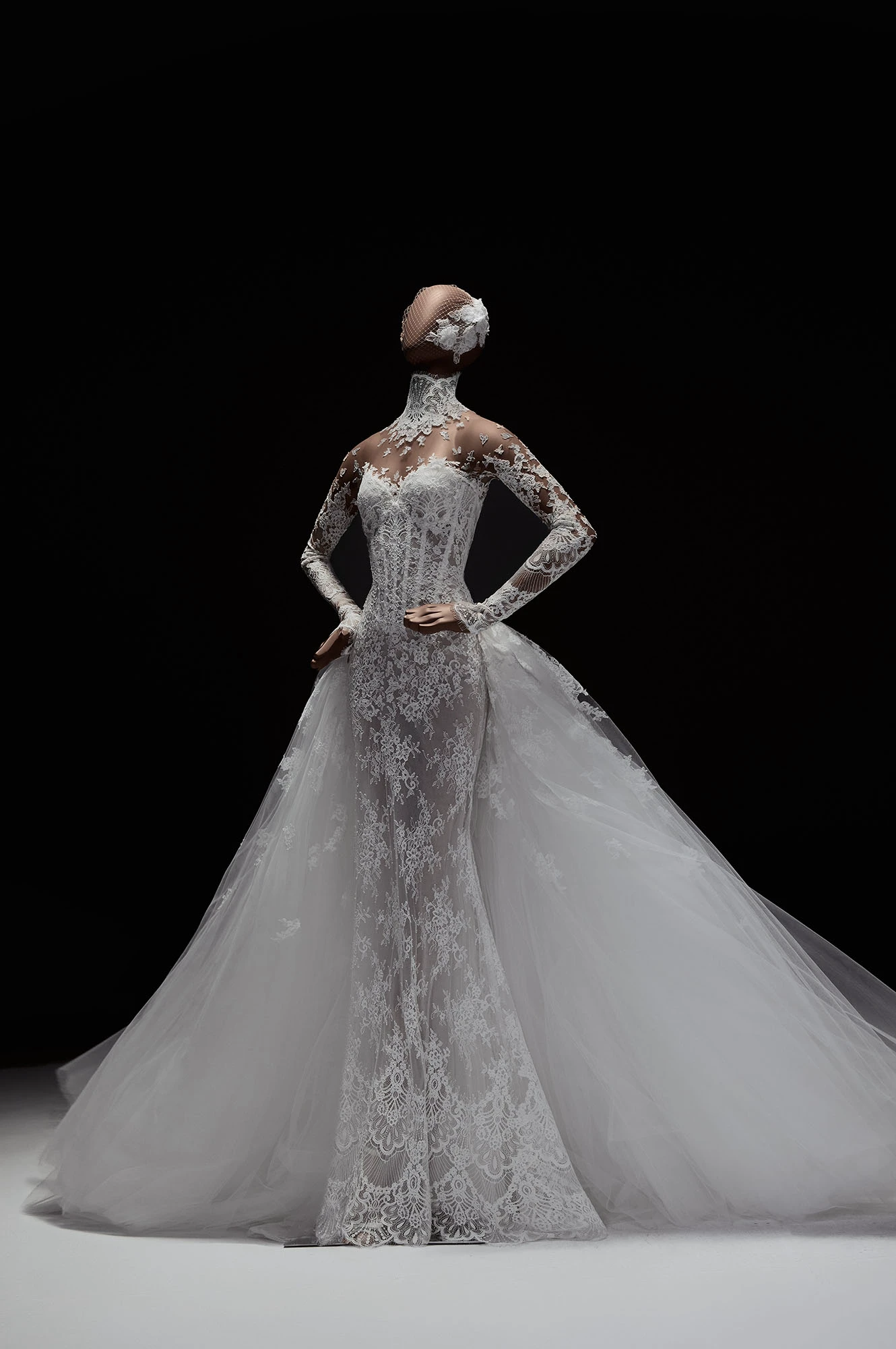 Elie Saab 2027 İlkbahar Gelinlik Koleksiyonu'ndan, Guipure dantel işlemeli, vücudu saran uzun kollu ve yüksek yakalı balık formlu bir gelinlik, dramatik ve kabarık tül kuyruğuyla masalsı bir siluet oluşturuyor, başındaki fileli peçe ve çiçek detaylarıyla zarif bir bütünlük sunuyor.