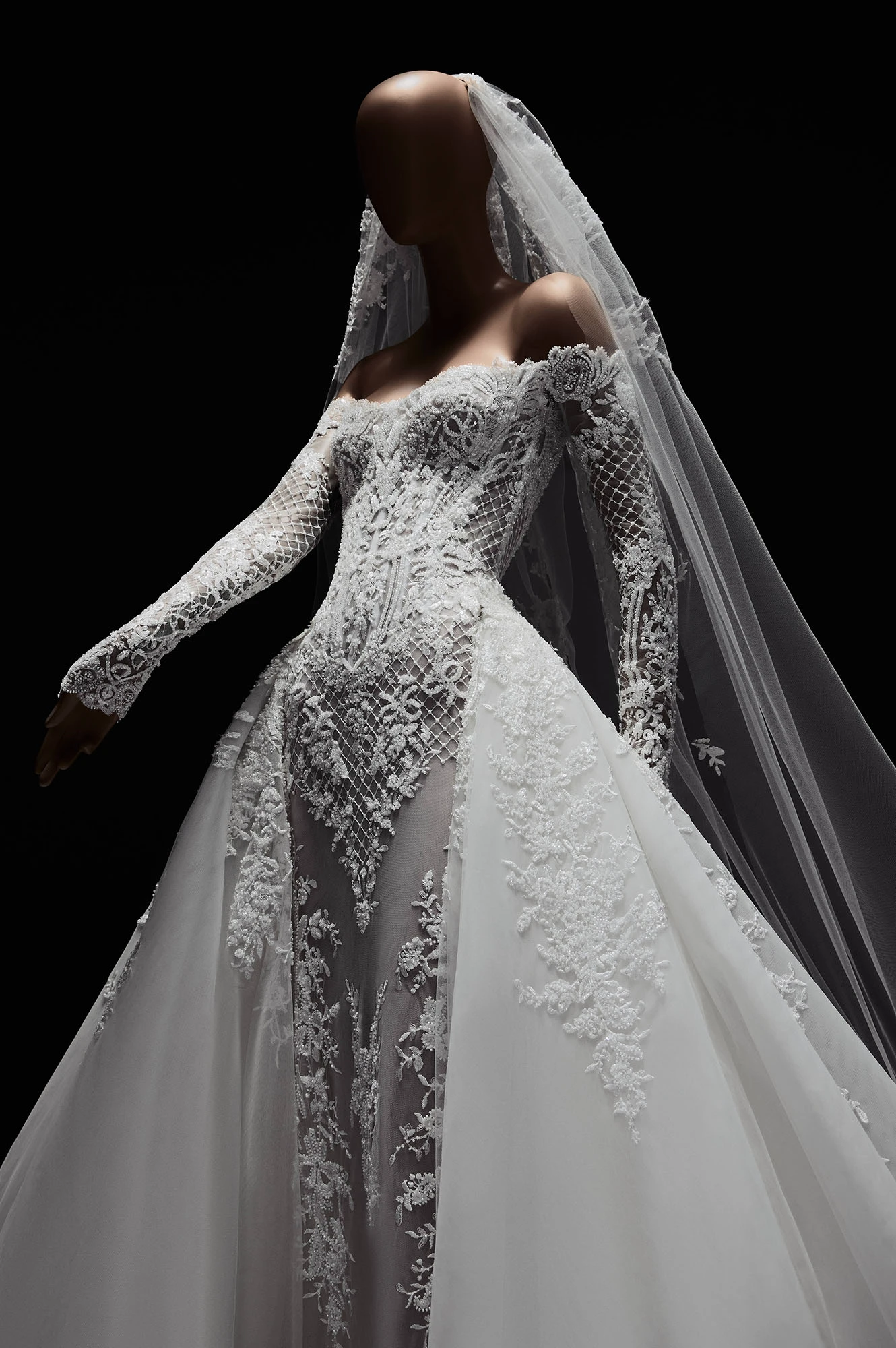 Mankendeki Elie Saab 2027 İlkbahar gelinlik koleksiyonundan bir parça olan bu zarif gelinlik, düşmüş omuzlu, dantel işlemeli uzun kolları, korse detaylı ve transparan filelerle süslenmiş üst kısmı, hacimli etekleri ve uzun işlemeli duvağıyla büyüleyici bir siluet sunuyor.