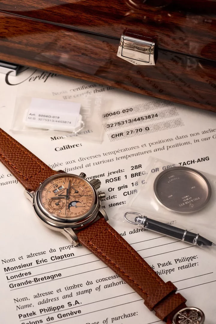 Somon renkli kadranı ve kahverengi deri kayışıyla dikkat çeken Patek Philippe perpetual takvim kronograf saati, Eric Clapton'ın adının geçtiği bir sertifika ile çeşitli yedek parçaların ve parlak ahşap bir kutunun önünde sergileniyor.