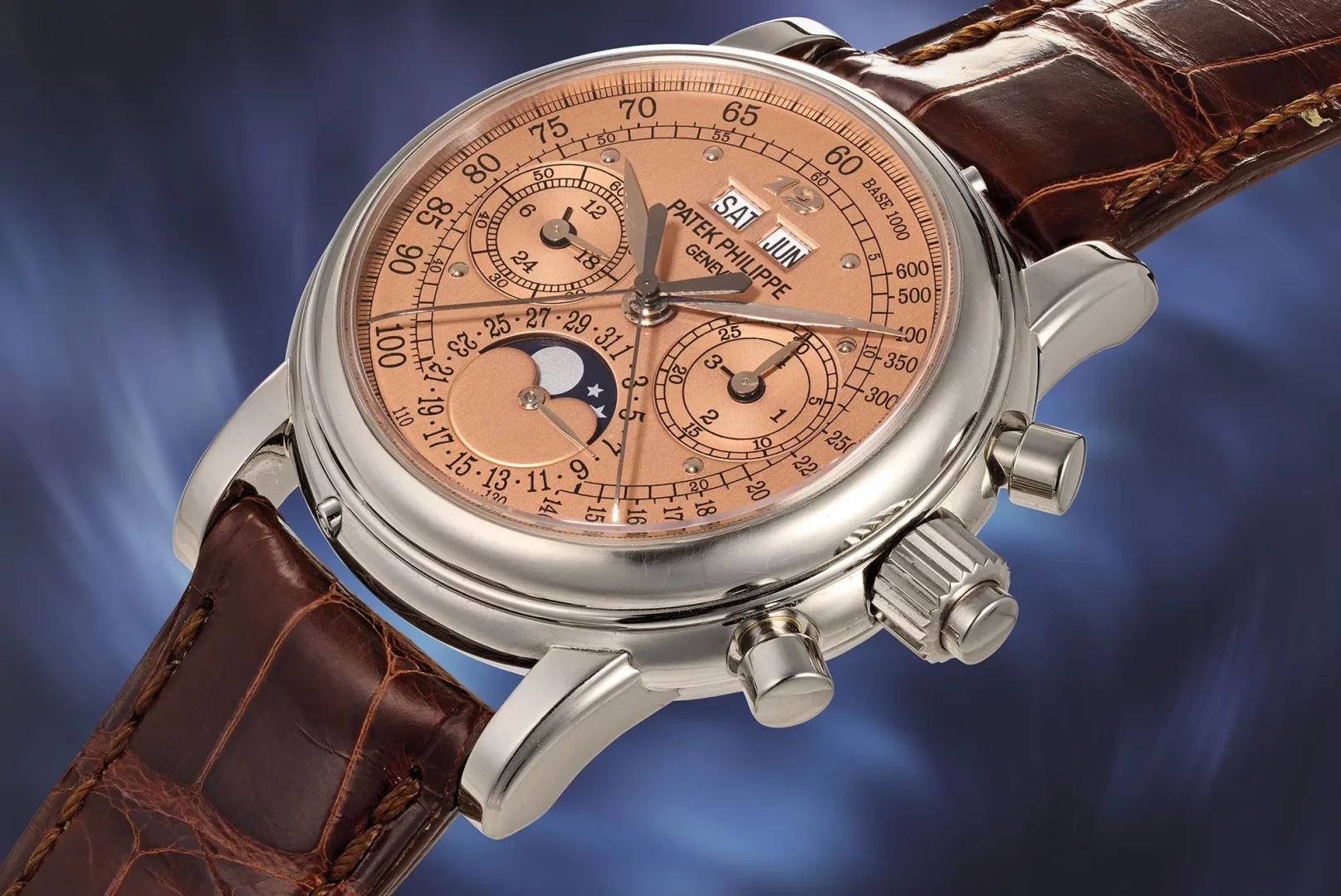 Patek Philippe'in somon rengi çok fonksiyonlu kadrana, ay fazı, gün, ay ve tarih göstergelerine sahip, parlak metal kasalı ve koyu kahverengi timsah derisi kayışlı lüks kronograf kol saati, ince işçiliğiyle dikkat çekiyor.