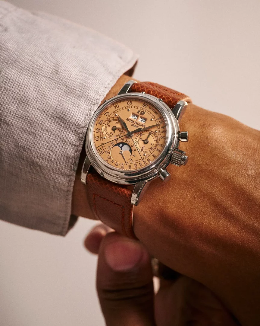 Açık gri renkli gömlek manşeti ve bronz tenli bir bilek üzerinde, Patek Philippe markalı, somon rengi çok fonksiyonlu kronograf kadranında takvim ve ay fazı göstergeleri barındıran, zarif gümüş kasalı ve kahverengi dokulu deri kayışlı lüks bir kol saati detaylı bir şekilde gösterilmektedir.